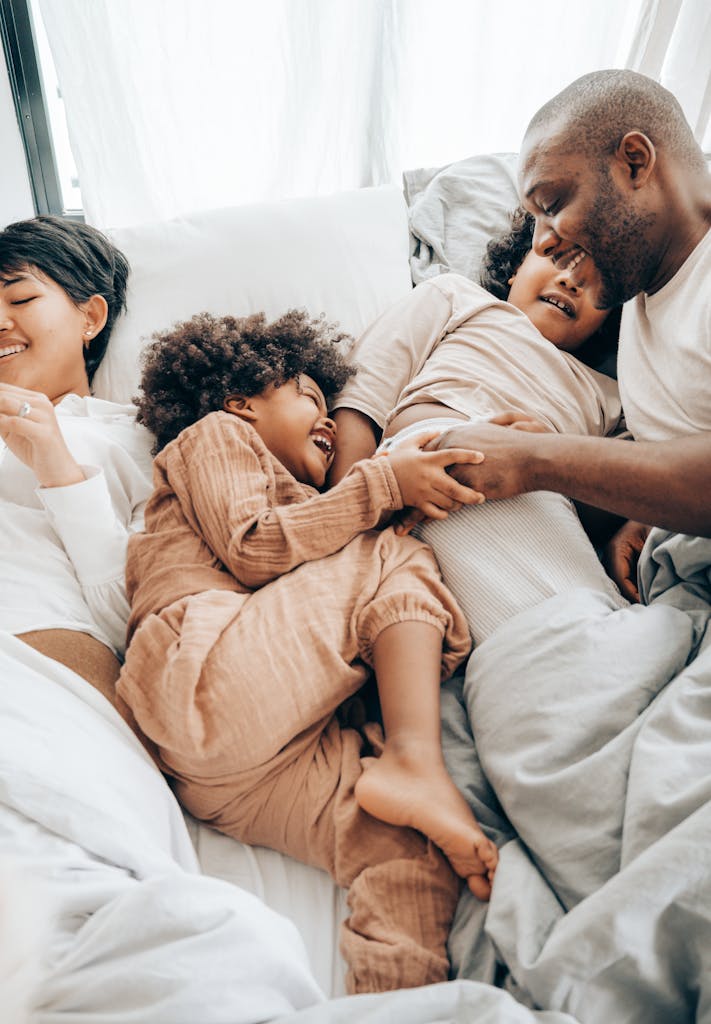 From above of smiling ethnic kids playing with father while resting together on bed lying near mother